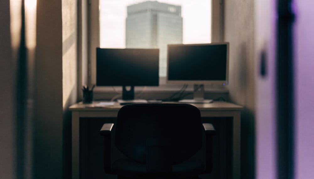 A quiet early morning view over a small office desk with two monitors showing lines of code, seen from behind an empty c