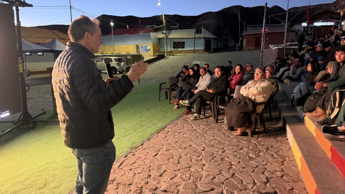 Un hombre con micrófono da una charla al aire libre, al anochecer, ante público sentado en sillas y graderías
