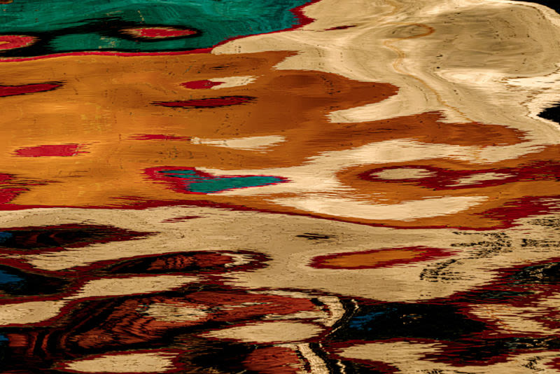 A graphic and energetic reinterpretation of the marine environment. The photograph focuses on the distorted and fragmented reflection of a colorful boat in the seawater. The water's surface acts as a natural lens, breaking down shapes and intensifying colors in an irregular and pulsating geometric pattern.