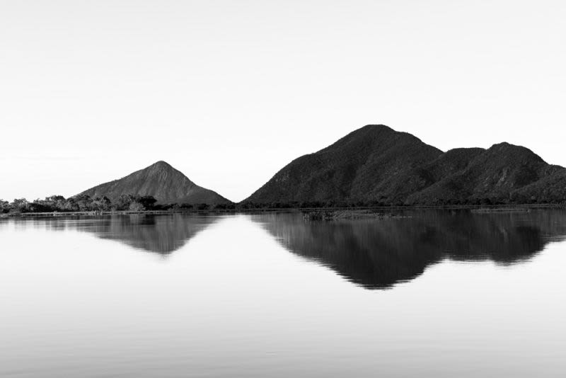 Una vista serena e intemporal de la emblemática sierra de Amolar en el Pantanal, capturada en un clásico y minimalista blanco y negro. La imagen plasma la majestuosa forma de la sierra, cuyos contornos se alzan abruptamente sobre el paisaje llano, reflejados con asombrosa claridad en un lago de perfecta calma.