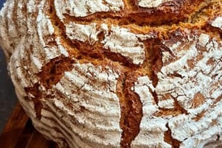 Rundes, goldbraunes Bauernbrot mit eingeschnittenem Muster, auf einem Holzbrett neben einer Schale Mehl