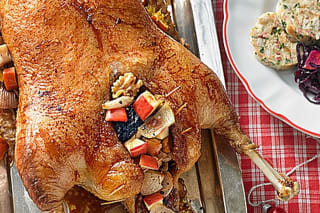 Serviettenknödel in Scheiben geschnitten, daneben goldbraune Gänsebrust und Rotkohl auf einem rustikalen Holzbrett