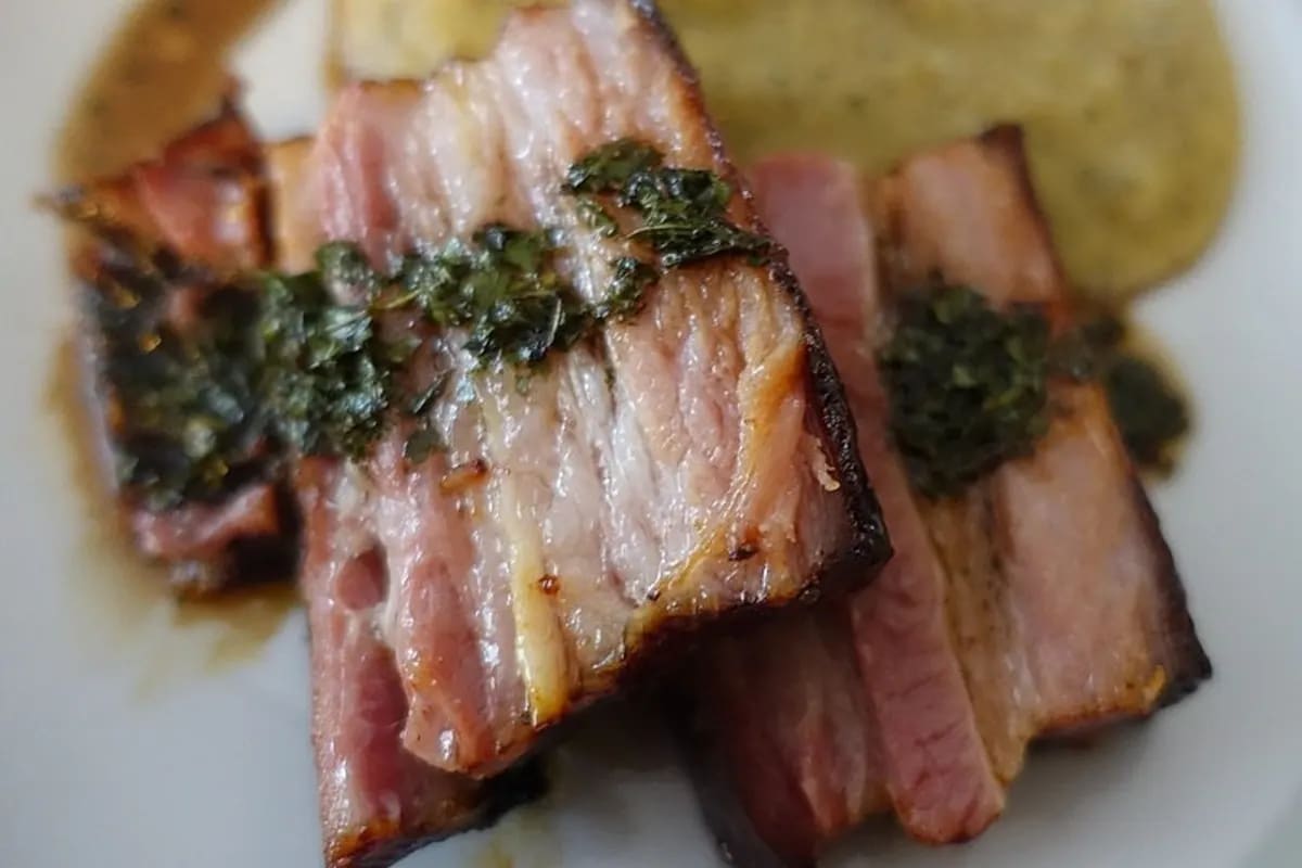 Honigglasierten Räucherspeck auf Holzbrett, daneben ein cremiges Minzspüree und ein Klecks grüner Minzsauce