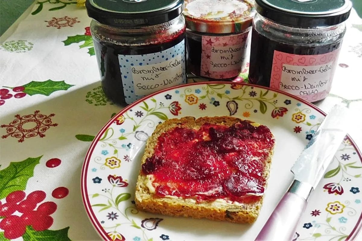 Gläser mit glänzendem Brombeergelee, daneben frische Brombeeren und eine Flasche Secco