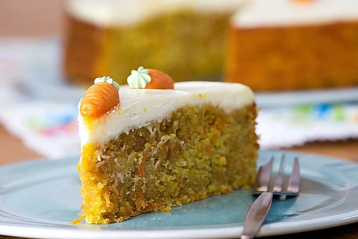 Goldbrauner Rüblikuchen mit glatter Frischkäse‑Zitronen‑Frosting, dekoriert mit geraspelten Karotten.