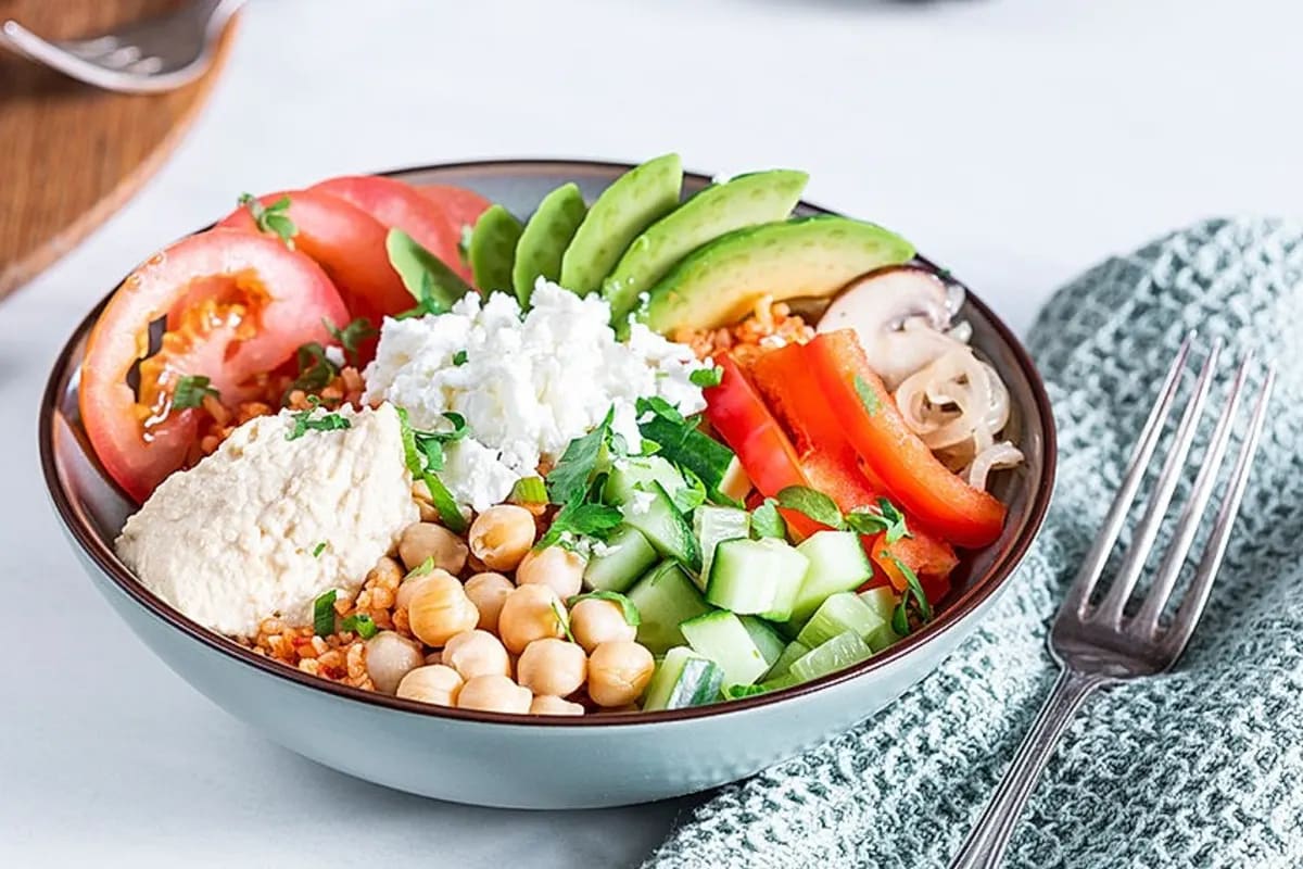 Bunte Buddha Bowl mit Bulgur, Paprika, Avocado, Pilzen, Kichererbsen und Hummus auf einem Holzbrett