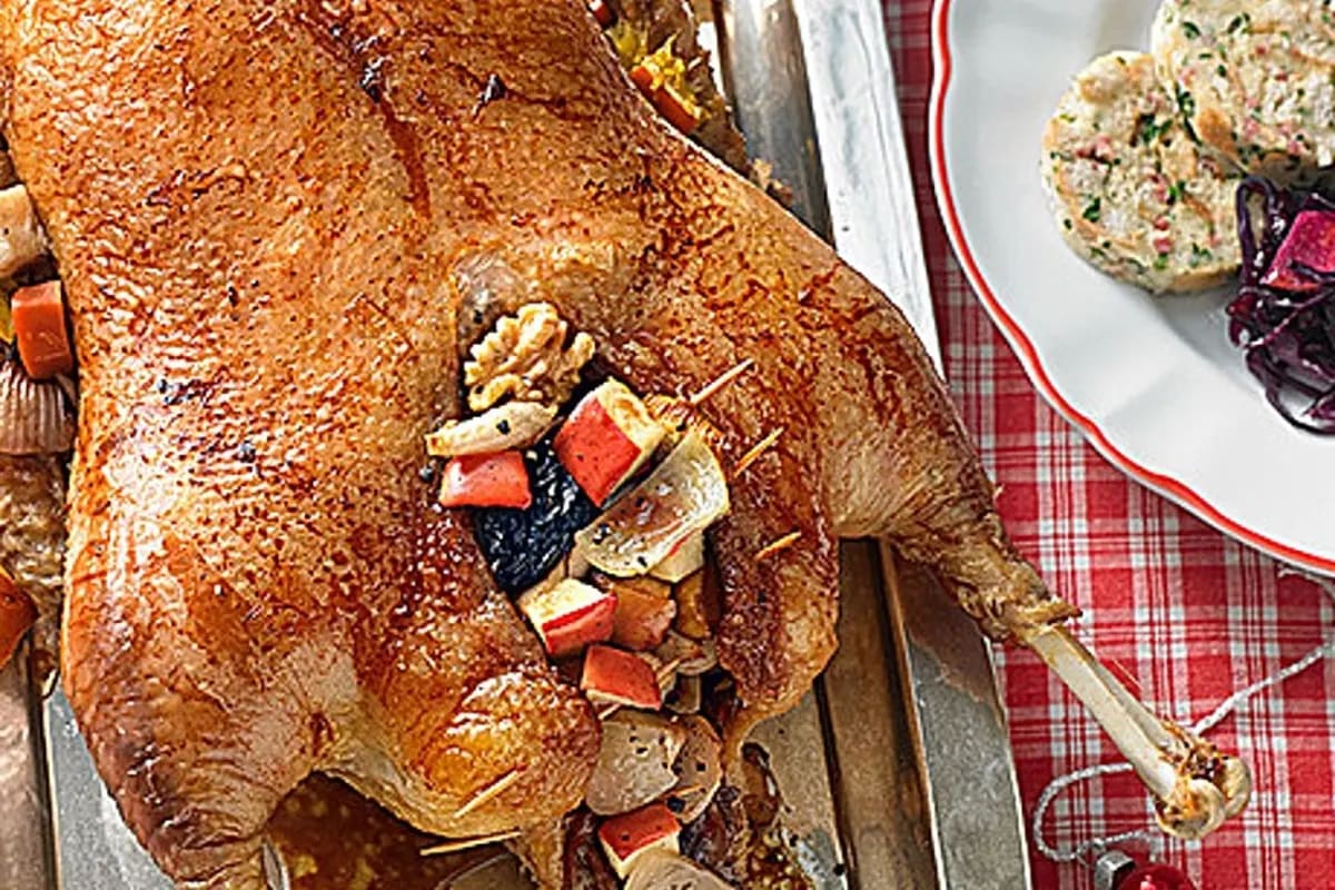 Serviettenknödel in Scheiben geschnitten, daneben goldbraune Gänsebrust und Rotkohl auf einem rustikalen Holzbrett