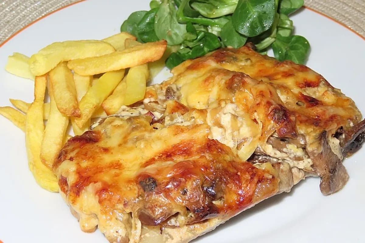 Goldbraun überbackene Schnitzel mit cremiger Pilz‑Sahnesauce, bestreut mit geschmolzenem Emmentaler, serviert neben einem Glas Weißwein und frischem Bauernbrot
