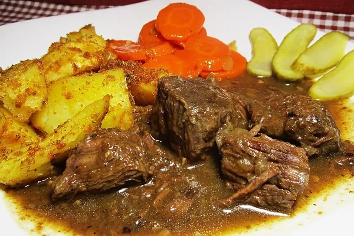 Dampfender Topf mit dunklem Pfefferpotthast, serviert neben Salzkartoffeln und Gewürzgurken