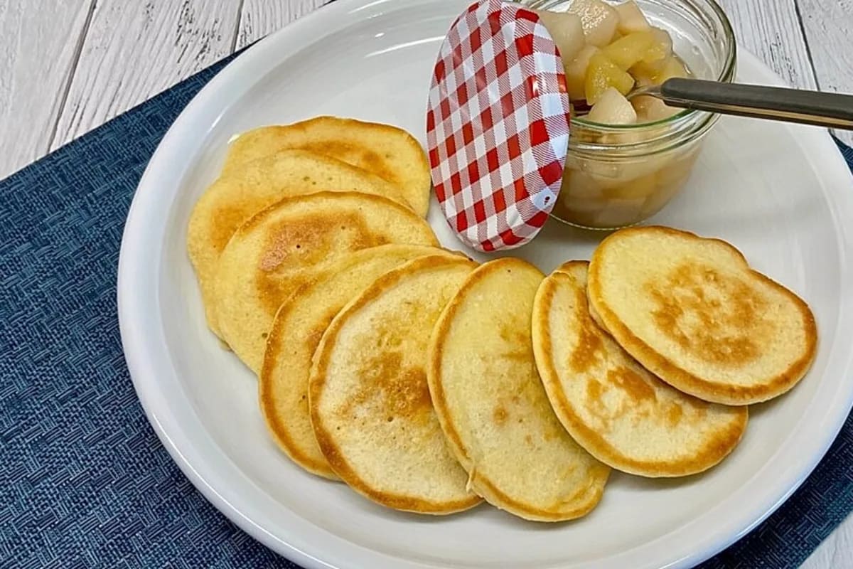 Kleine goldbraune Pfannkuchen, gestapelt auf einem Teller, bestäubt mit Puderzucker und einem Klecks Apfelmus