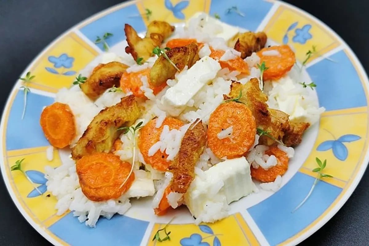 Bunter saftiger Reissalat mit goldenen Möhren, saftigen Hühnchenstücken, Rosinen und zerbröckeltem Feta auf einem weißen Teller