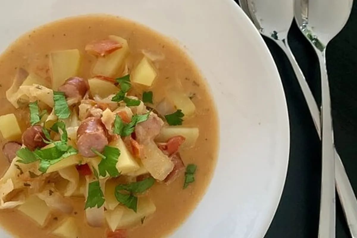 Bunter Eintopf aus Chinakohl, Kartoffeln und Debreziner-Würstchen, serviert in einem Topf mit frischem Sauerrahm