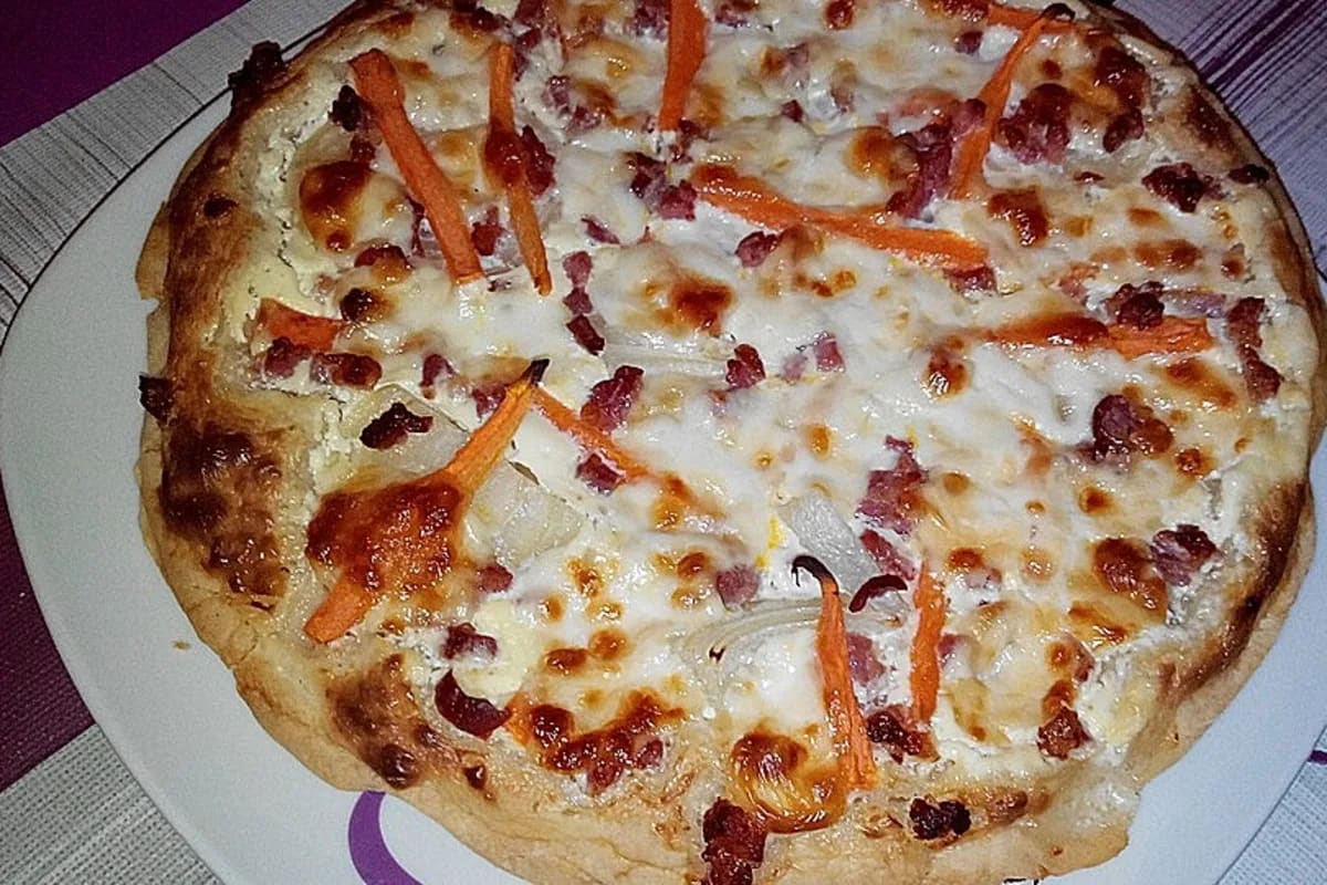 Bunter Flammkuchen mit Zwiebelringen, Möhrenstreifen, geschmolzenem Mozzarella und knusprigem Speck auf Holzbrett