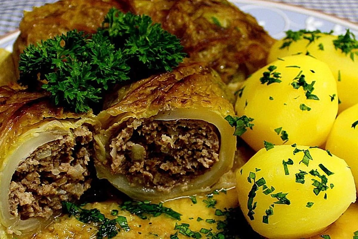 Goldbraun gebratene Kohlrouladen liegen in einer cremigen Sauce, daneben dampfende Salzkartoffeln auf einem rustikalen Holzteller