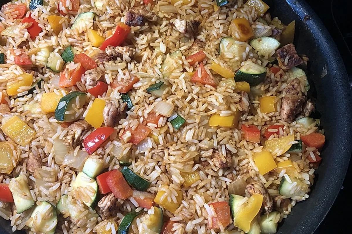 Pfanne mit knusprigem Reis, goldbrauner Putenstreifen, roter Paprika und grüner Zucchini, angerichtet auf Holzbrett