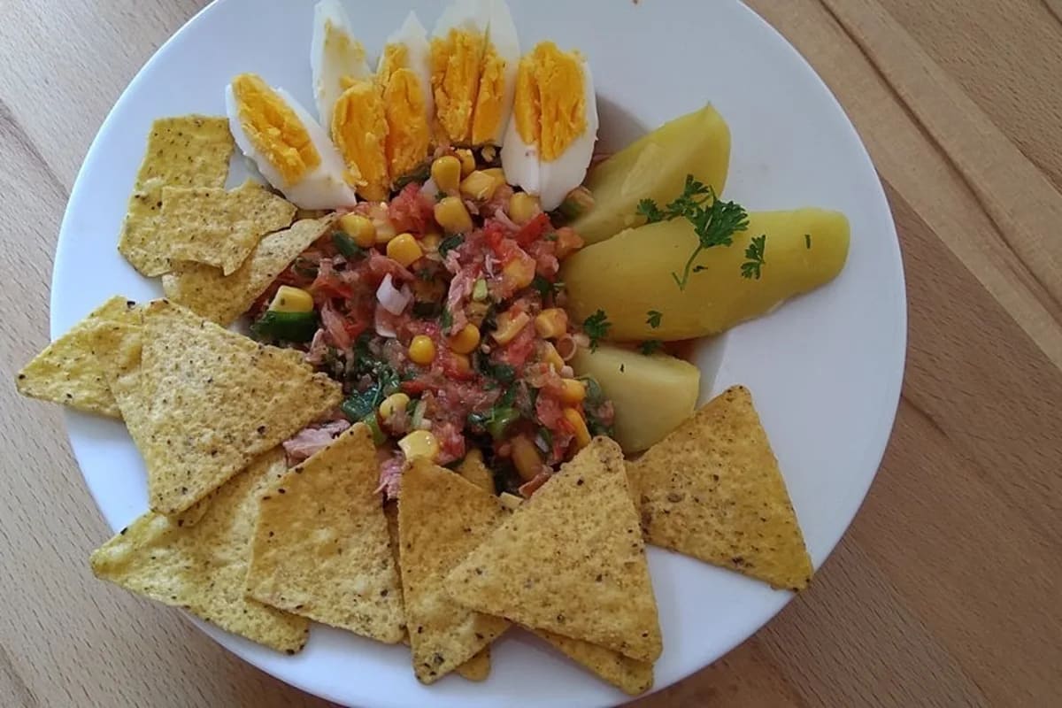 Kalte Tomatencreme mit Thunfisch, Mais, geviertelten Eiern und knusprigen Nachos, dazu daneben servierte Kartoffelviertel auf rustikalem Holzbrett