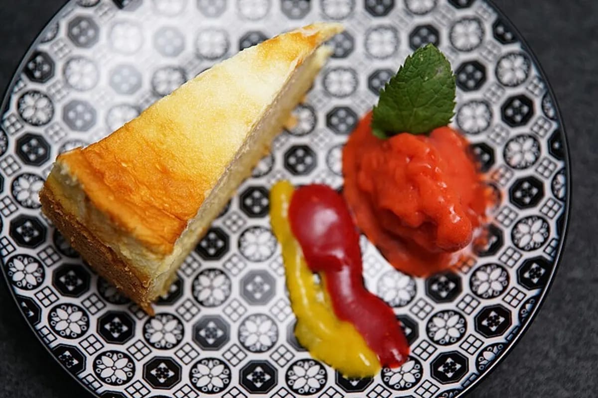 Zwei‑schichtiger Käsekuchen mit goldbraunem Baiser, Mango‑Himbeer‑Sorbet und leuchtendem Fruchtspiegel auf weißem Teller