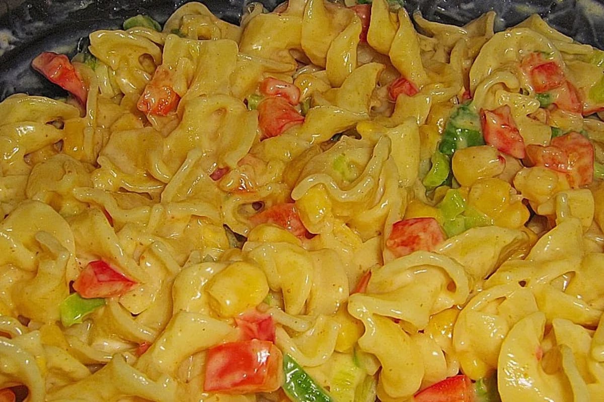 Bunter Nudelsalat mit gelbem Curry‑Dressing, roten Paprikawürfeln, grünen Porreeringen und frischer Petersilie auf einem Holzbrett