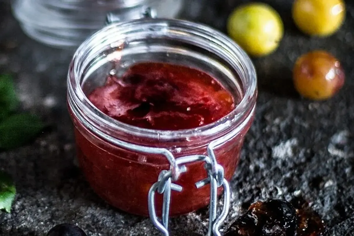 Glas mit roter, glänzender Mirabellenmarmelade, Löffel daneben, rustikale Holztheke im Hintergrund