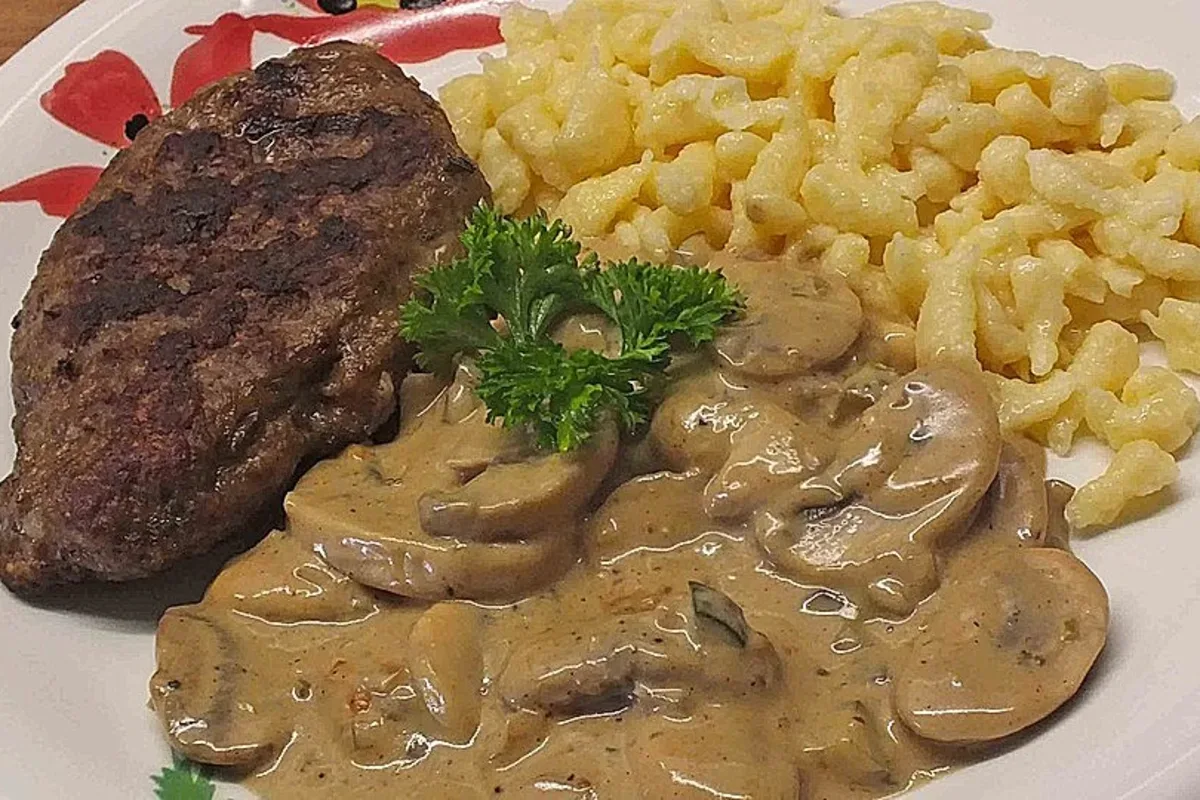 Goldbraun gebratene Hacksteaks auf einem Teller neben cremiger Pilz‑Sahne‑Sauce und Nudeln