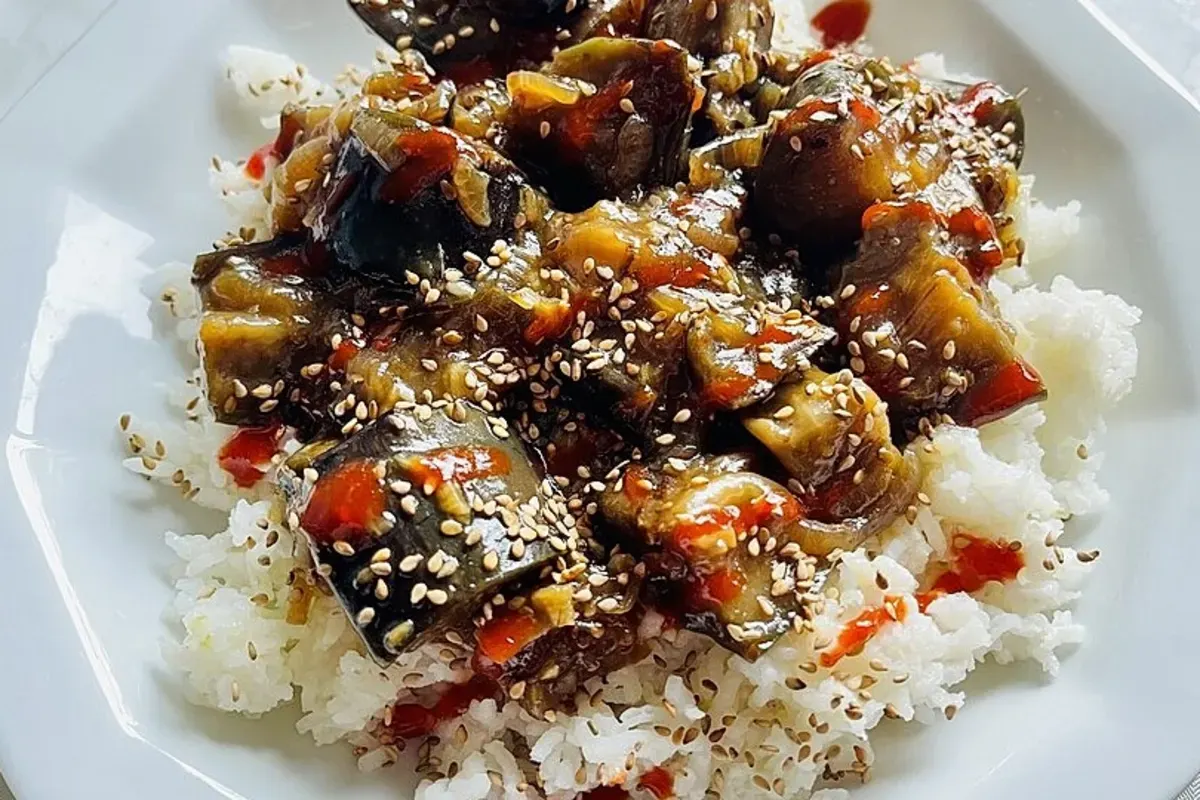 Gebratene Auberginenstücke in einer wok‑geeigneten Pfanne, glasiert mit dunkler Sojasaucen‑Reduktion, garniert mit Chili und Sesamkörnern.