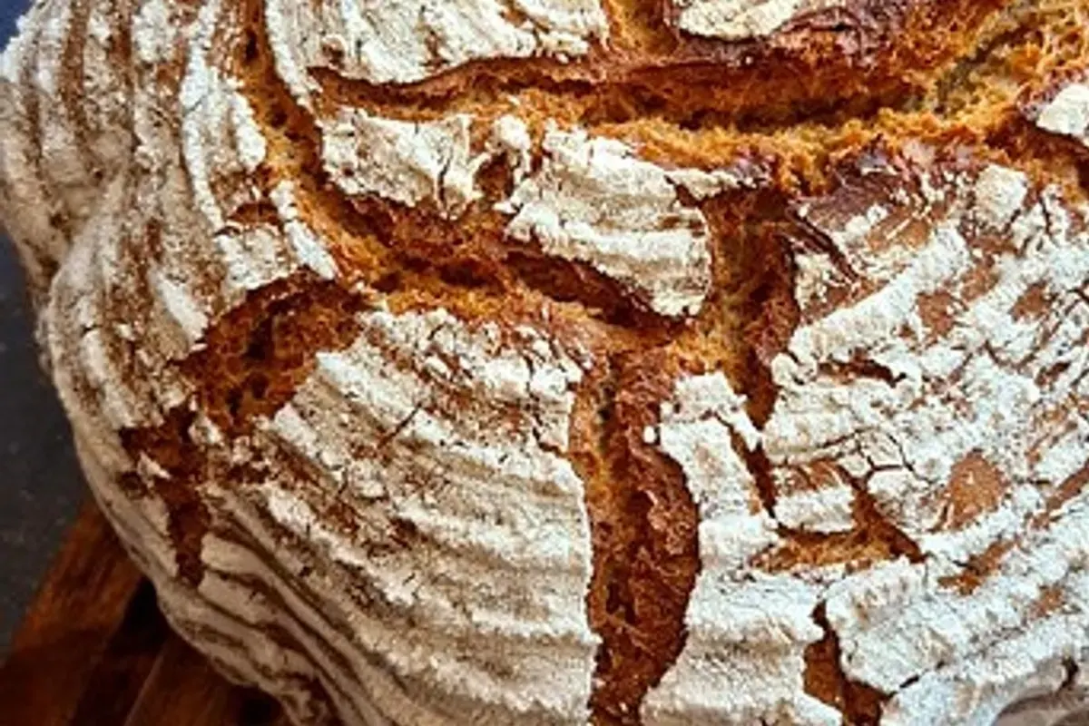 Rundes, goldbraunes Bauernbrot mit eingeschnittenem Muster, auf einem Holzbrett neben einer Schale Mehl