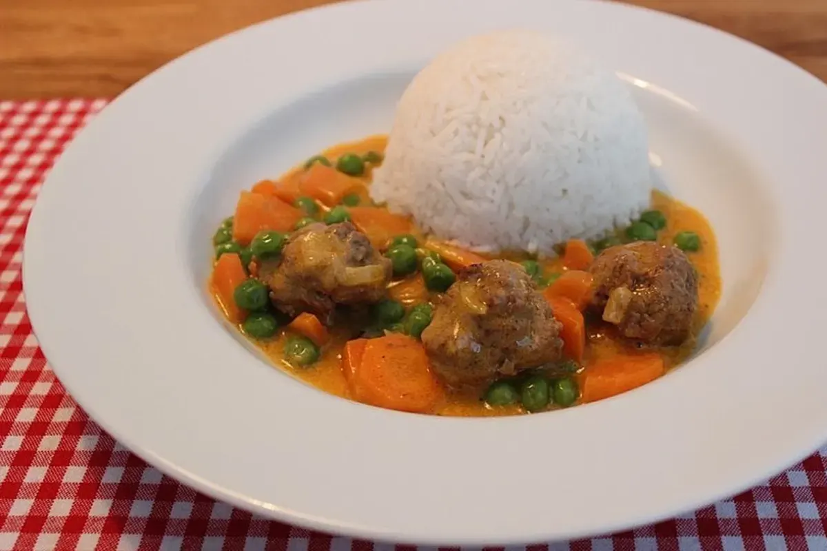 Großer Topf mit goldbraunen Hackbällchen, grünen Erbsen, orangefarbenen Möhren und cremiger Sauce, garniert mit frischer Petersilie