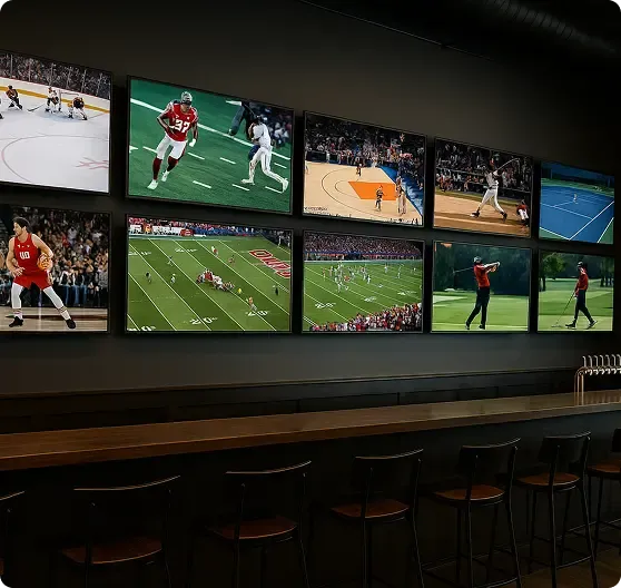 A row of TV screens above a bar shows various live sports including football, basketball, baseball, tennis, and golf; empty barstools line the counter.
