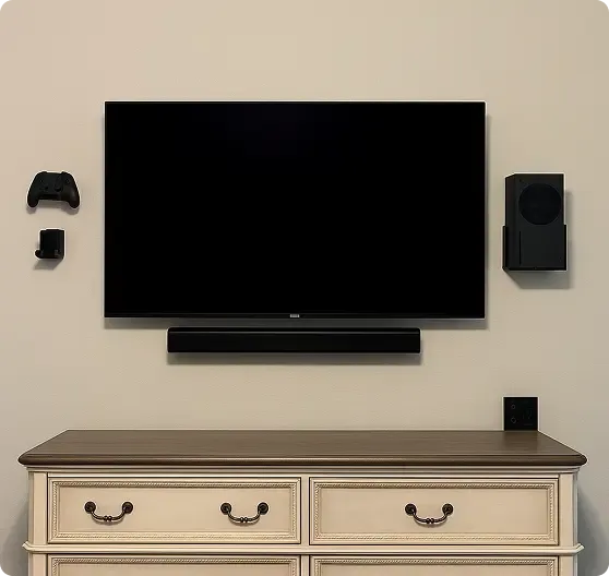A wall-mounted flatscreen TV with a soundbar below, flanked by a game controller, remote, and speaker, above a cream-colored dresser with four drawers.