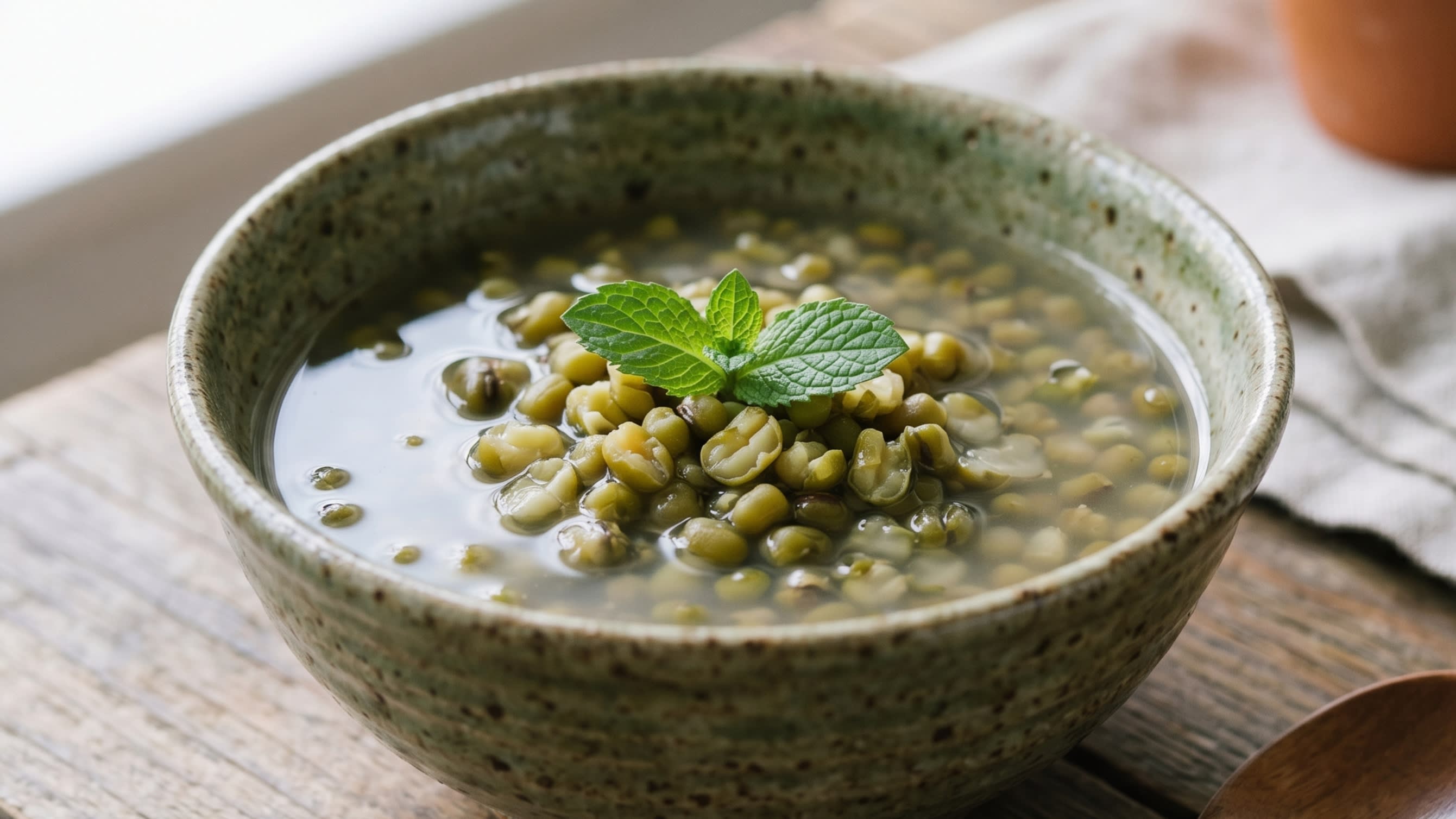 A close-up shot of a rustic ceramic bowl filled with sweet mung bean soup, the green beans perfectly split open, garnished with a single mint leaf (16:9 ratio)