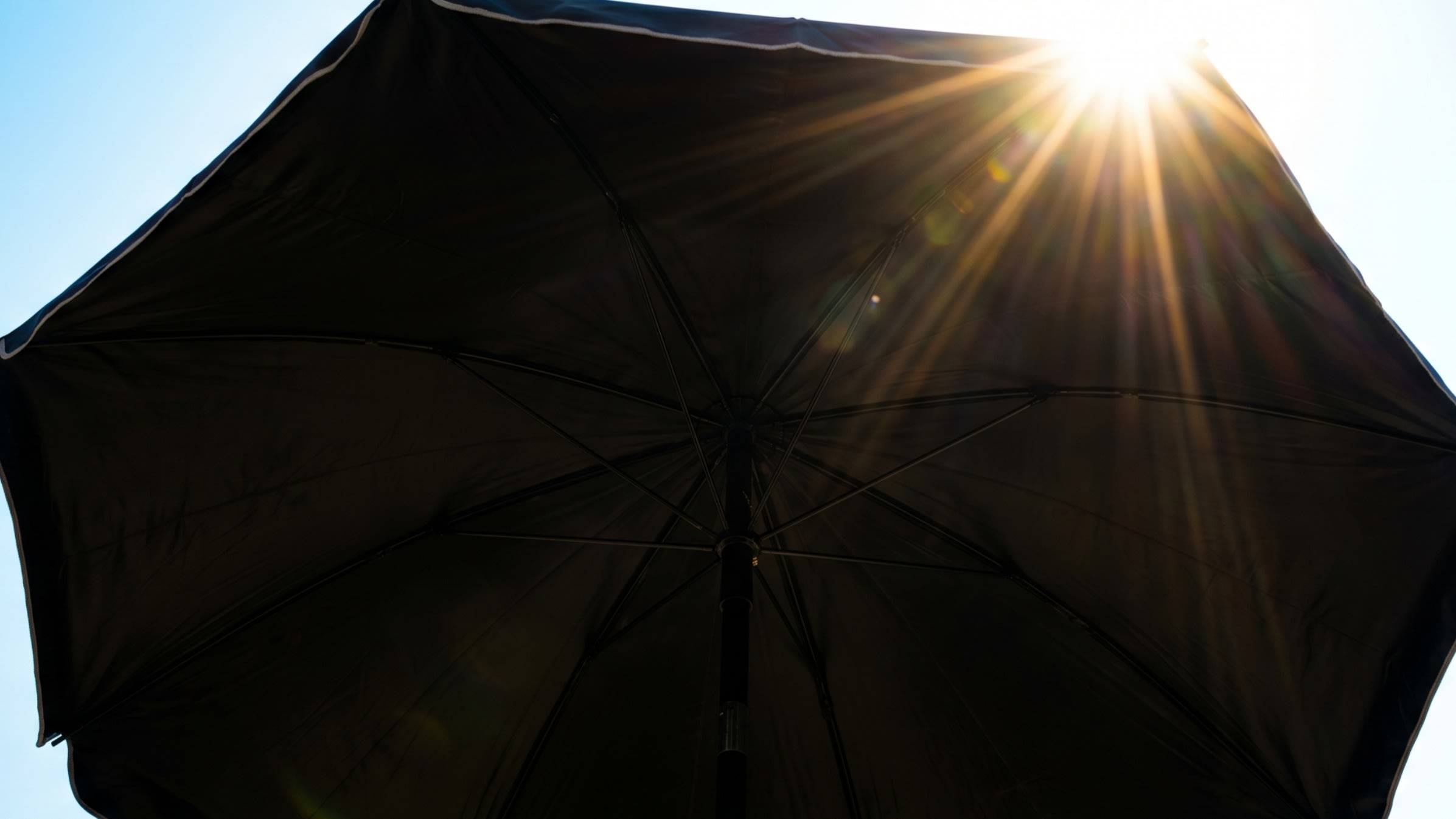 A close-up shot from beneath a black-coated parasol, looking up at the blinding sun. The thick black vinyl completely blocks the light, creating a stark contrast of deep shadow inside the umbrella (16:9 ratio)