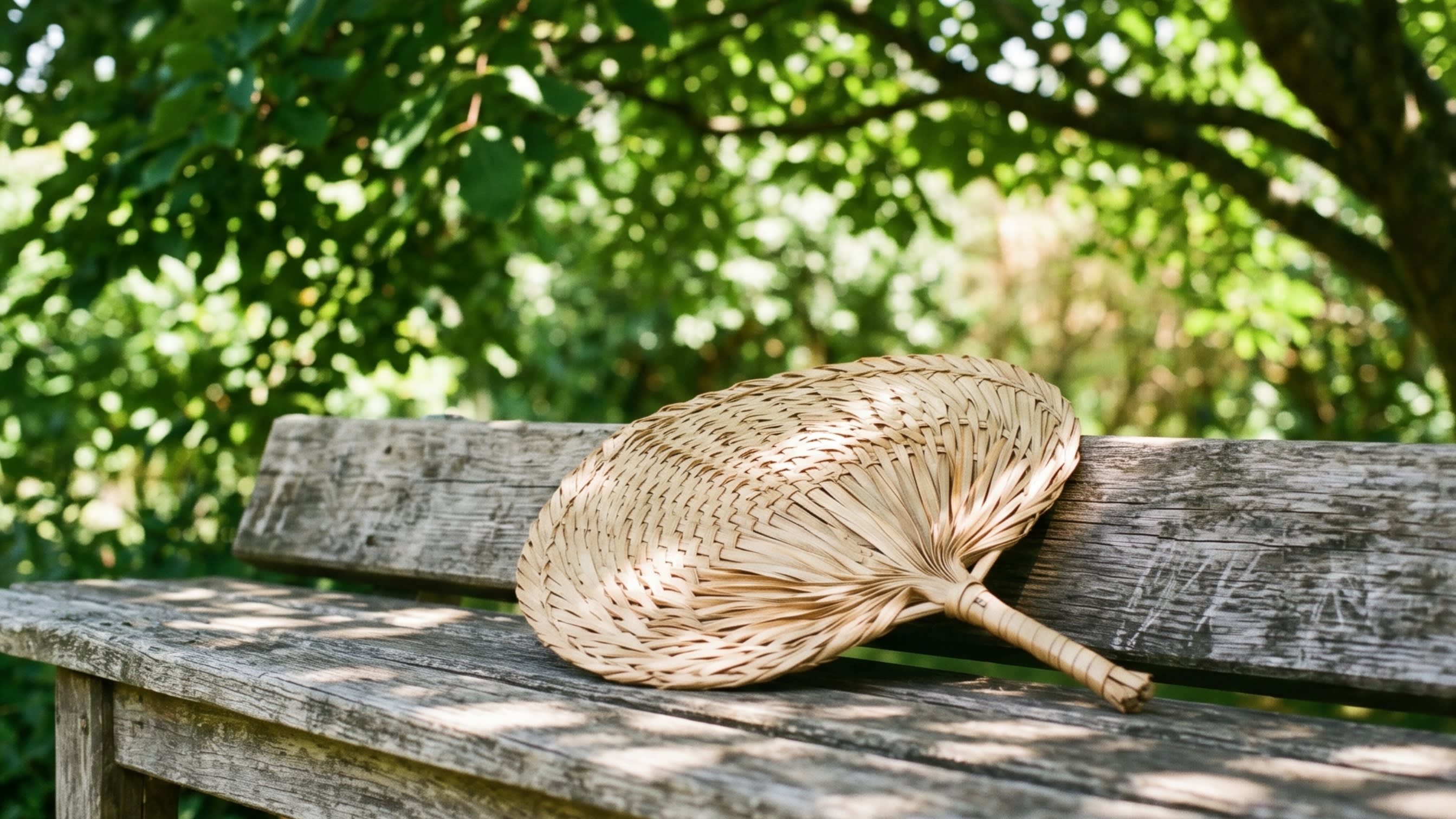 A close-up shot of a rustic, hand-woven palm-leaf fan resting on a vintage wooden bench under the dappled shade of a large tree (16:9 ratio)
