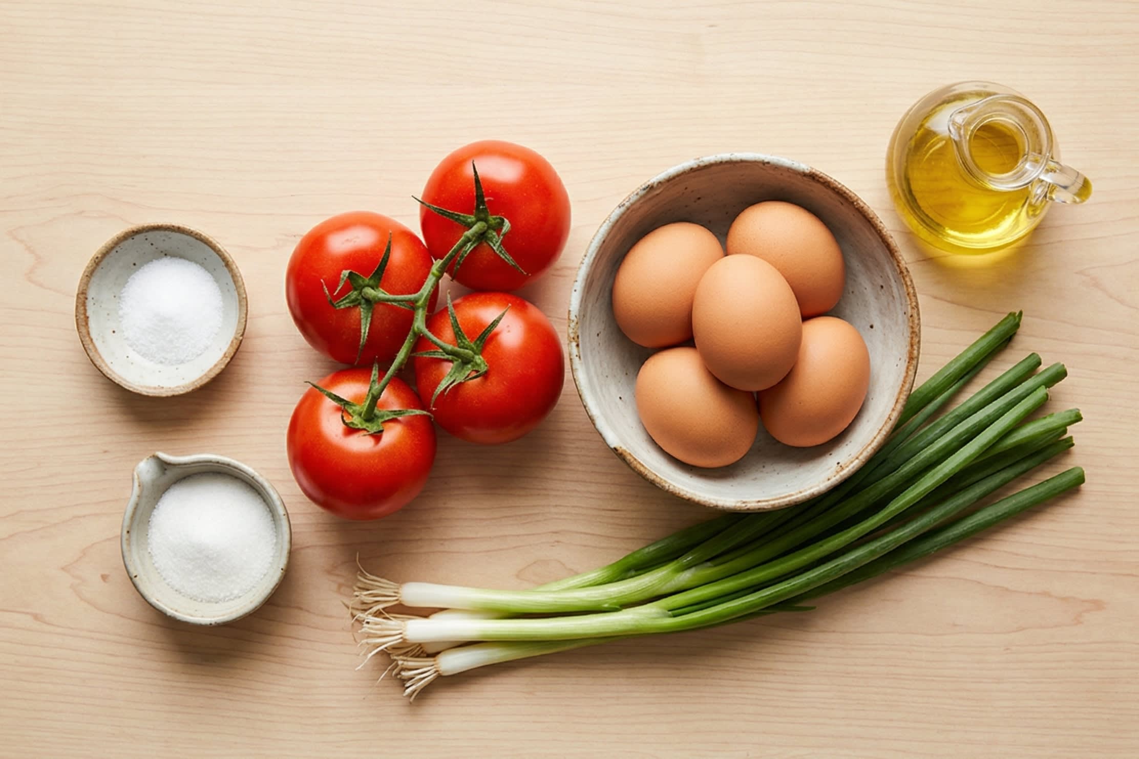 A bright, top-down flat lay showing the simple, vibrant ingredients: four ripe red tomatoes on the vine, four large brown eggs, fresh green scallions, and small ceramic dishes containing salt, sugar, and cooking oil