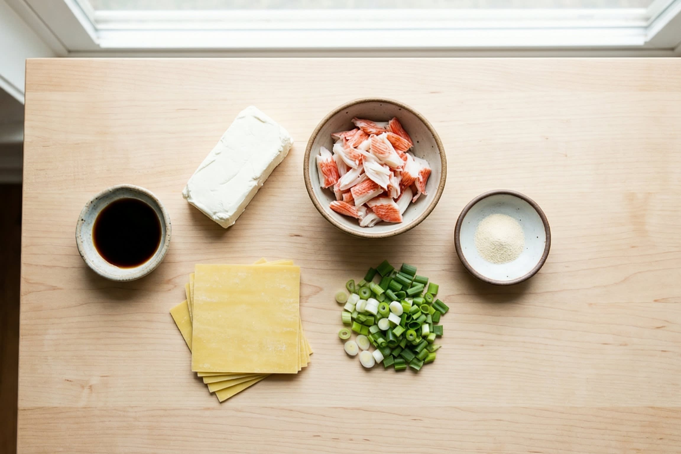 A vibrant, top-down flat lay food photography shot showing the raw ingredients: a block of cream cheese, a package of square wonton wrappers, shredded imitation crab meat, fresh scallions, and small dipping bowls of soy sauce, Worcestershire sauce, and garlic powder.
