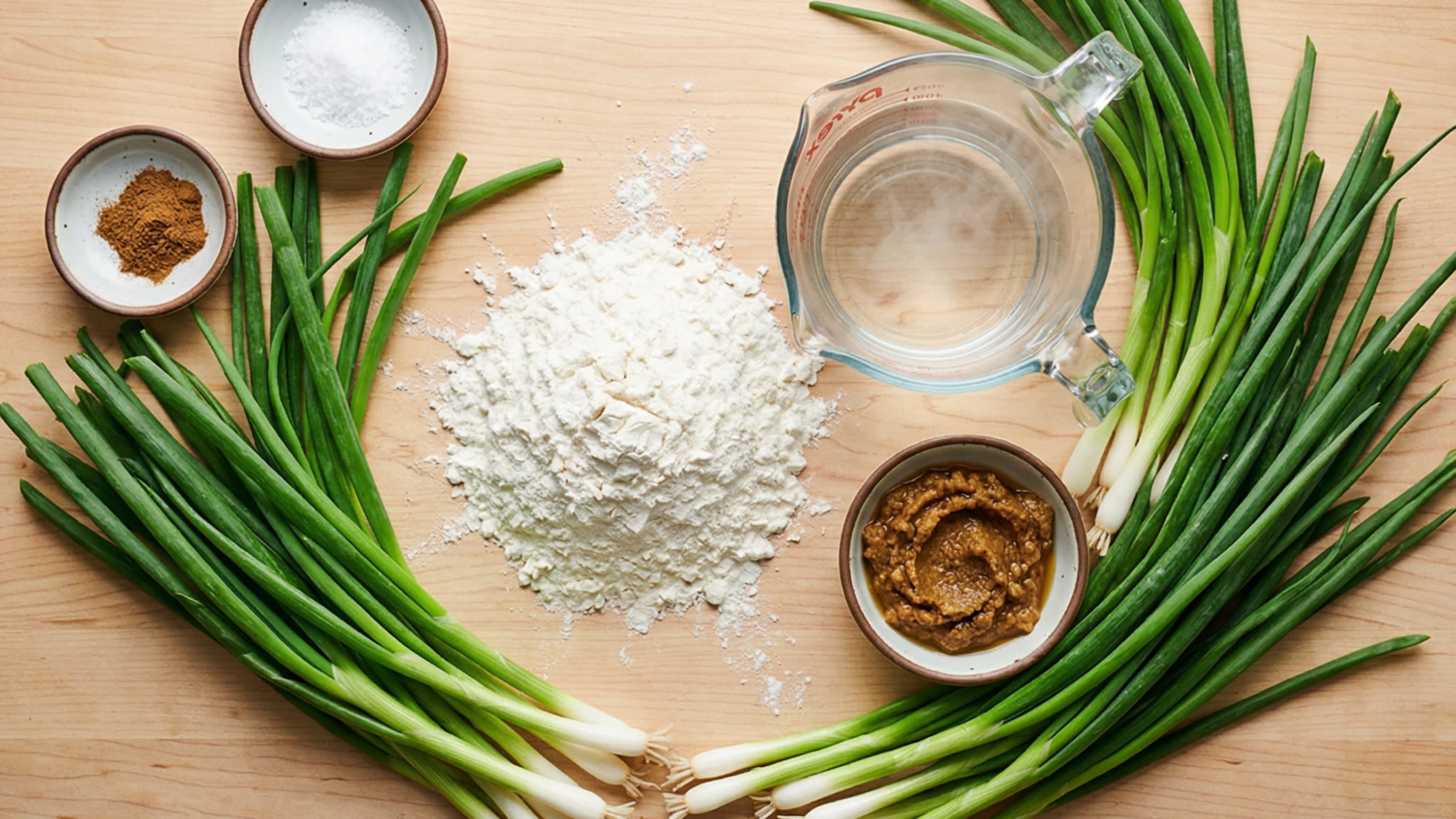 A beautiful flat lay of fresh ingredients on a wooden board: a mound of all-purpose flour, a glass measuring cup of boiling water, a massive bunch of fresh, vibrant green scallions, and small ceramic bowls containing salt, five-spice powder, and a thick, savory oil roux (Yousu).