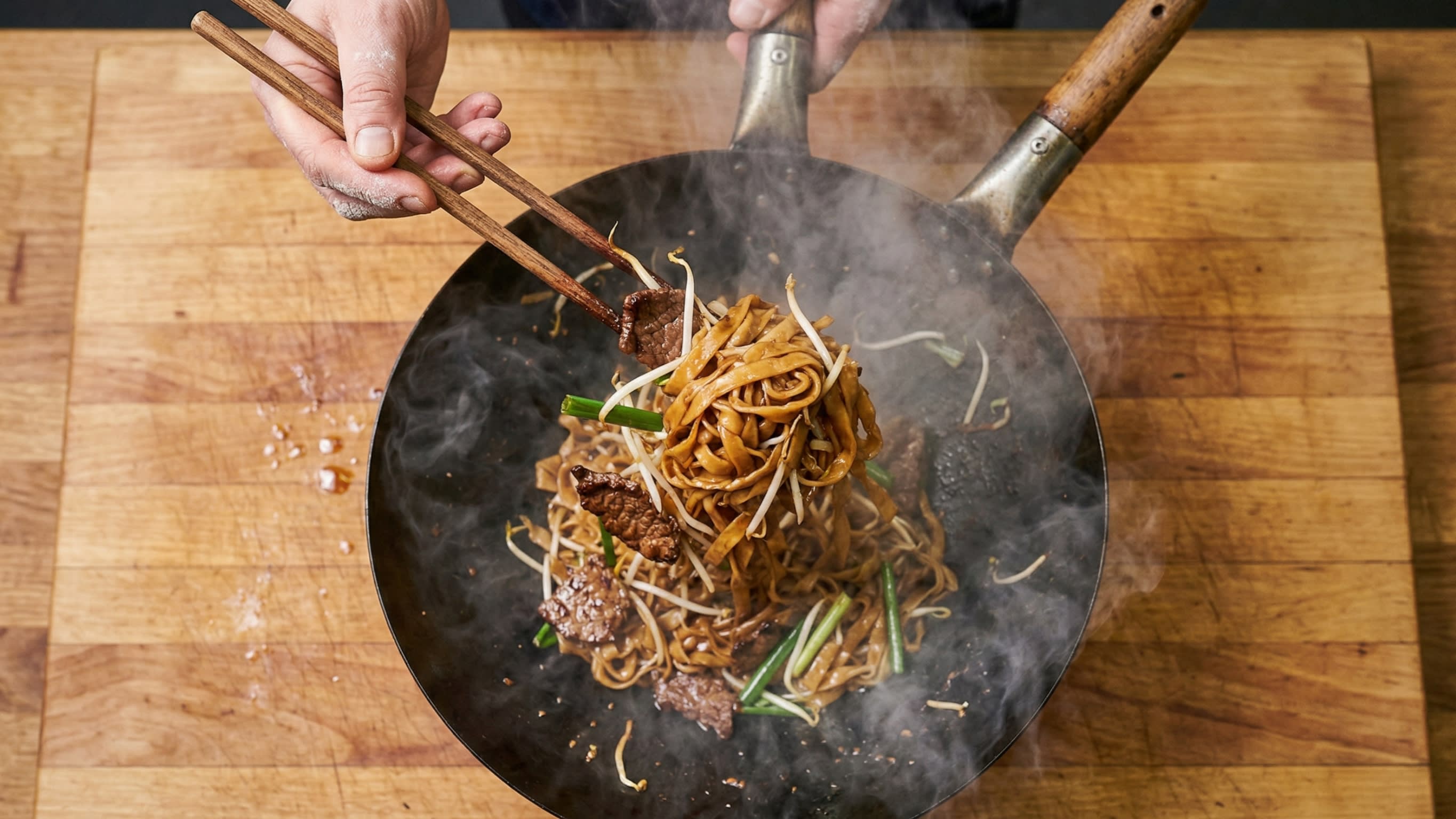 A dynamic action food photography shot looking down into a hot, well-seasoned traditional carbon steel wok over a bright roaring flame. A chef is using extra-long wooden cooking chopsticks to aggressively toss and lift a tangle of wide, dark mahogany rice noodles. Hot plumes of appetizing steam and "Wok Hei" smoke are rising.