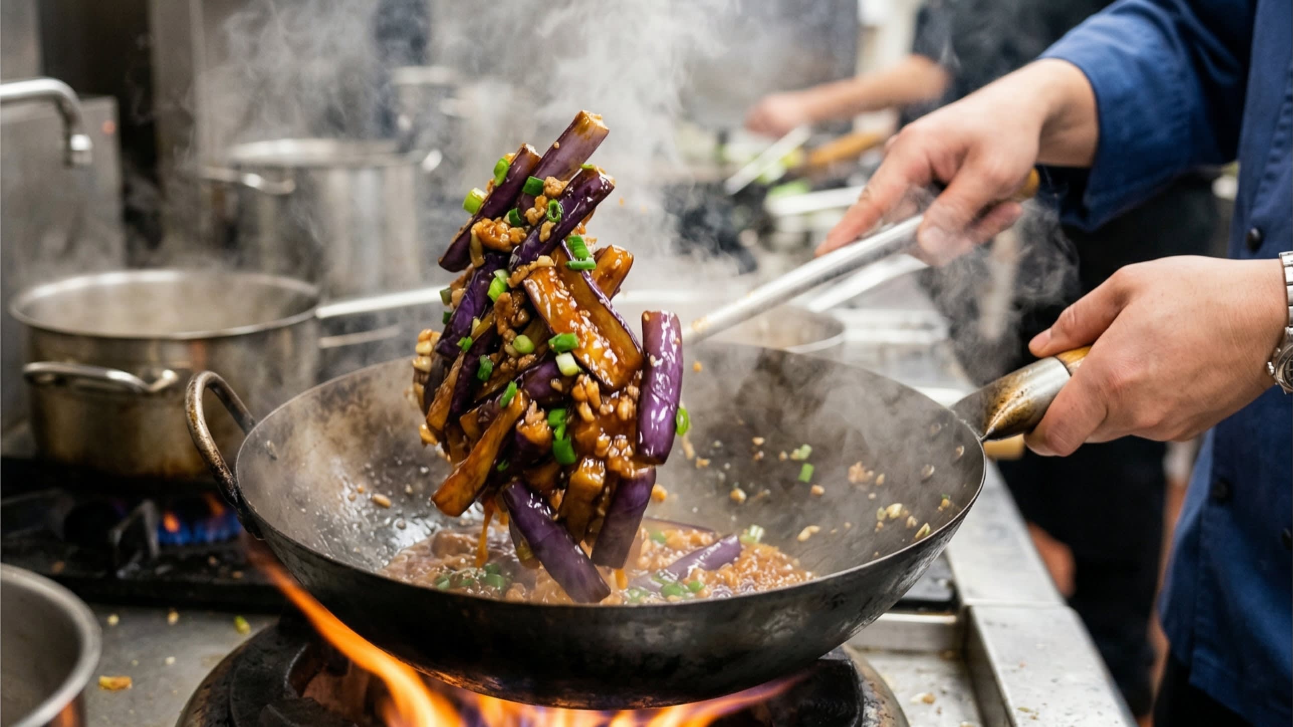 A vibrant action food photography shot looking down into a hot, traditional carbon steel wok over a bright flame. A chef is aggressively tossing the velvety, soft, golden-brown eggplant batons with a thick, glossy, bubbling, reddish-brown garlic sauce. Bright green scallions are mixed in, and hot steam is rising.