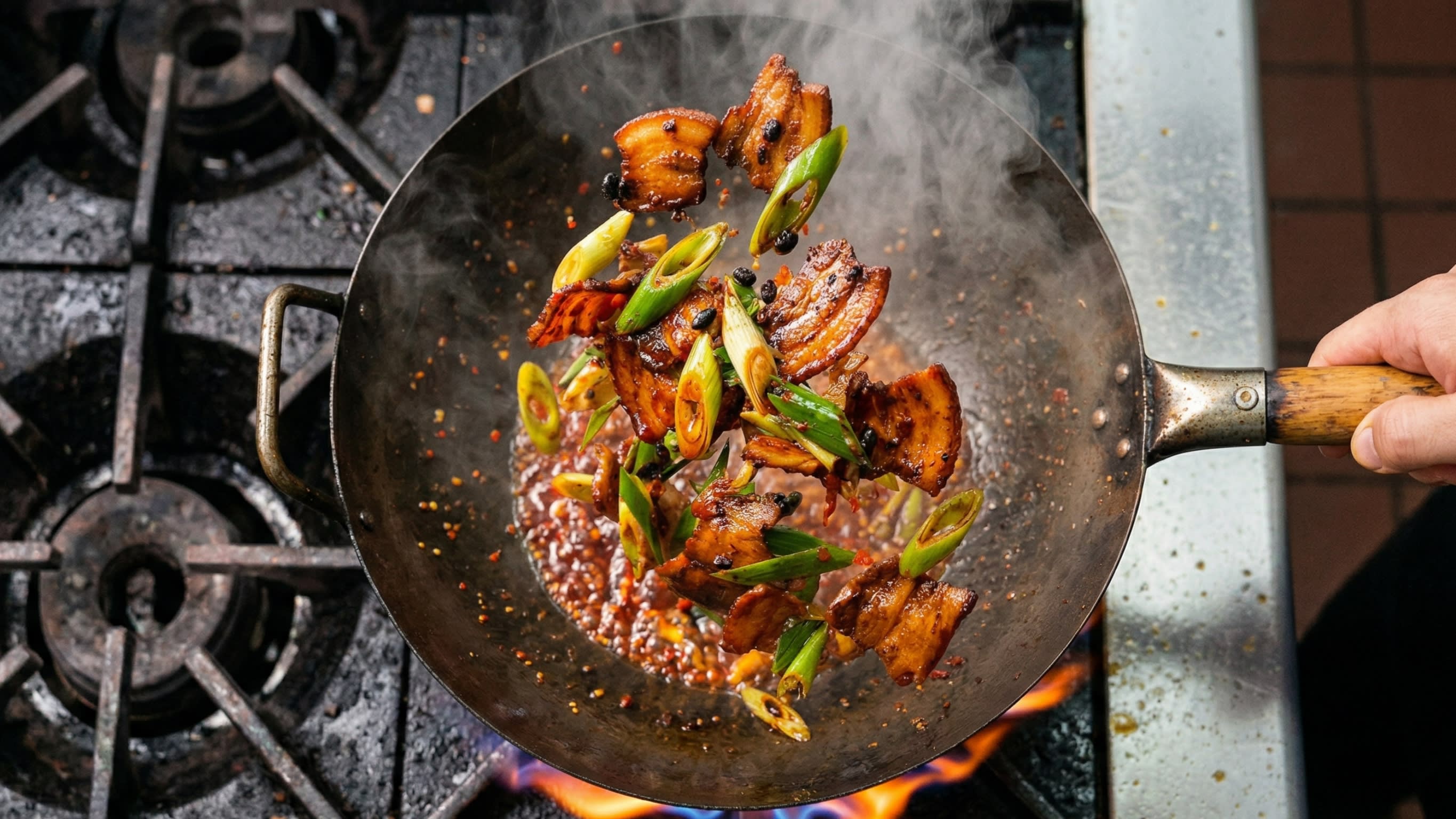 A vibrant action food photography shot looking directly down into a hot, traditional carbon steel wok over a bright flame. A chef is aggressively tossing the beautifully curled, caramelized pork belly slices with a thick, glossy, bubbling, fiery ruby-red sauce and bright green, diagonally sliced leeks. Hot steam is rising dramatically.
