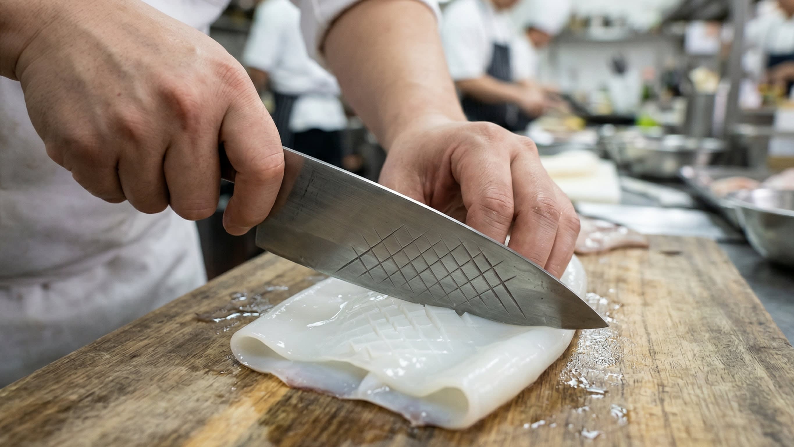 A dynamic close-up action shot focusing on a chef's hands executing the "Pinecone" cut. A sharp, traditional Chinese cleaver is delicately scoring a tight, diagonal crosshatch pattern onto the inside surface of a flattened, translucent white squid tube on a wooden cutting board.