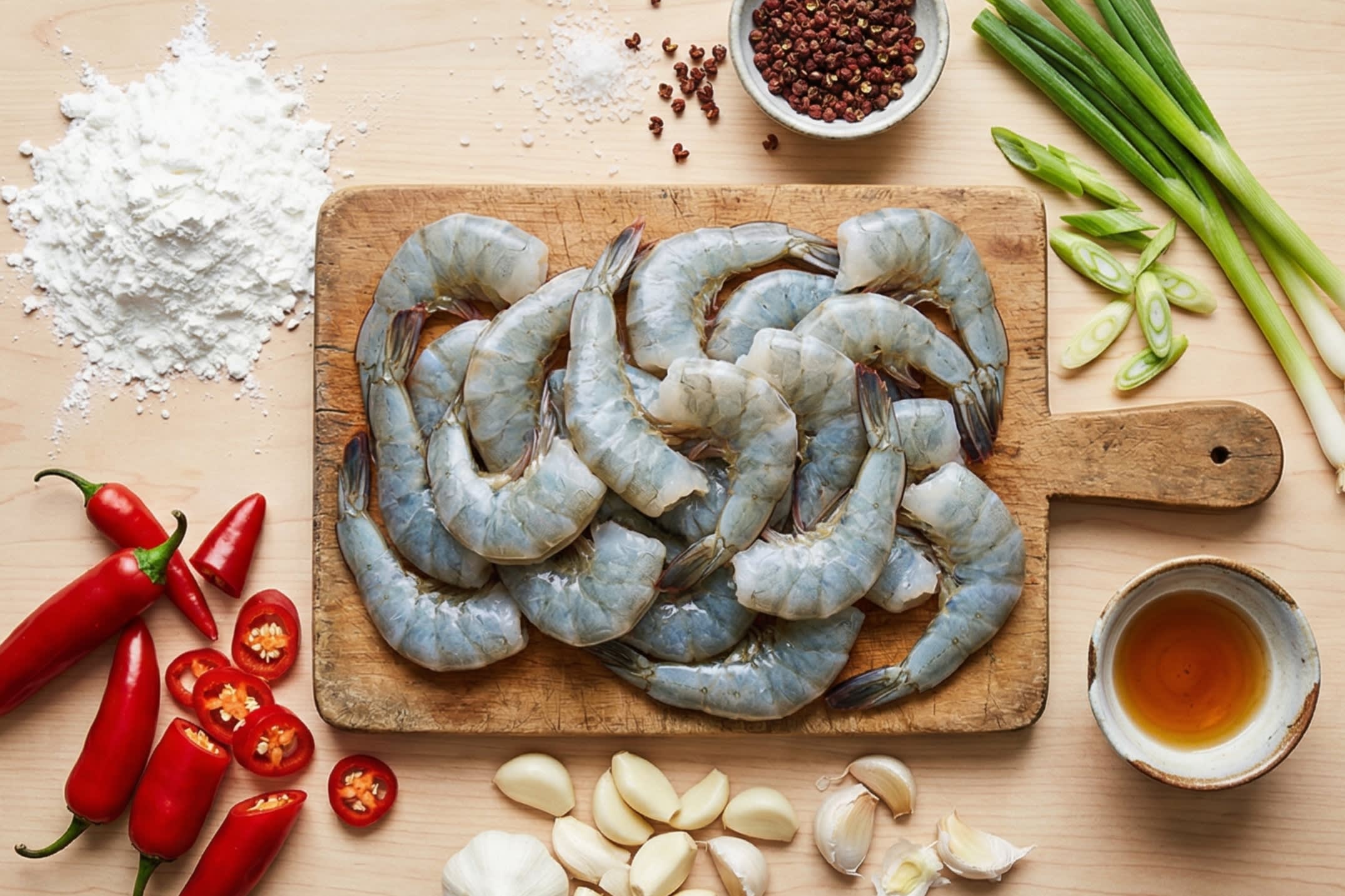 A beautiful flat lay of fresh ingredients on a wooden board: large, raw shell-on shrimp with a deep slit cut down their backs, a mound of pure white cornstarch, fiery red jalapeño peppers, fresh garlic cloves, vibrant green scallions, whole Sichuan peppercorns, coarse sea salt crystals, and Shaoxing cooking wine.