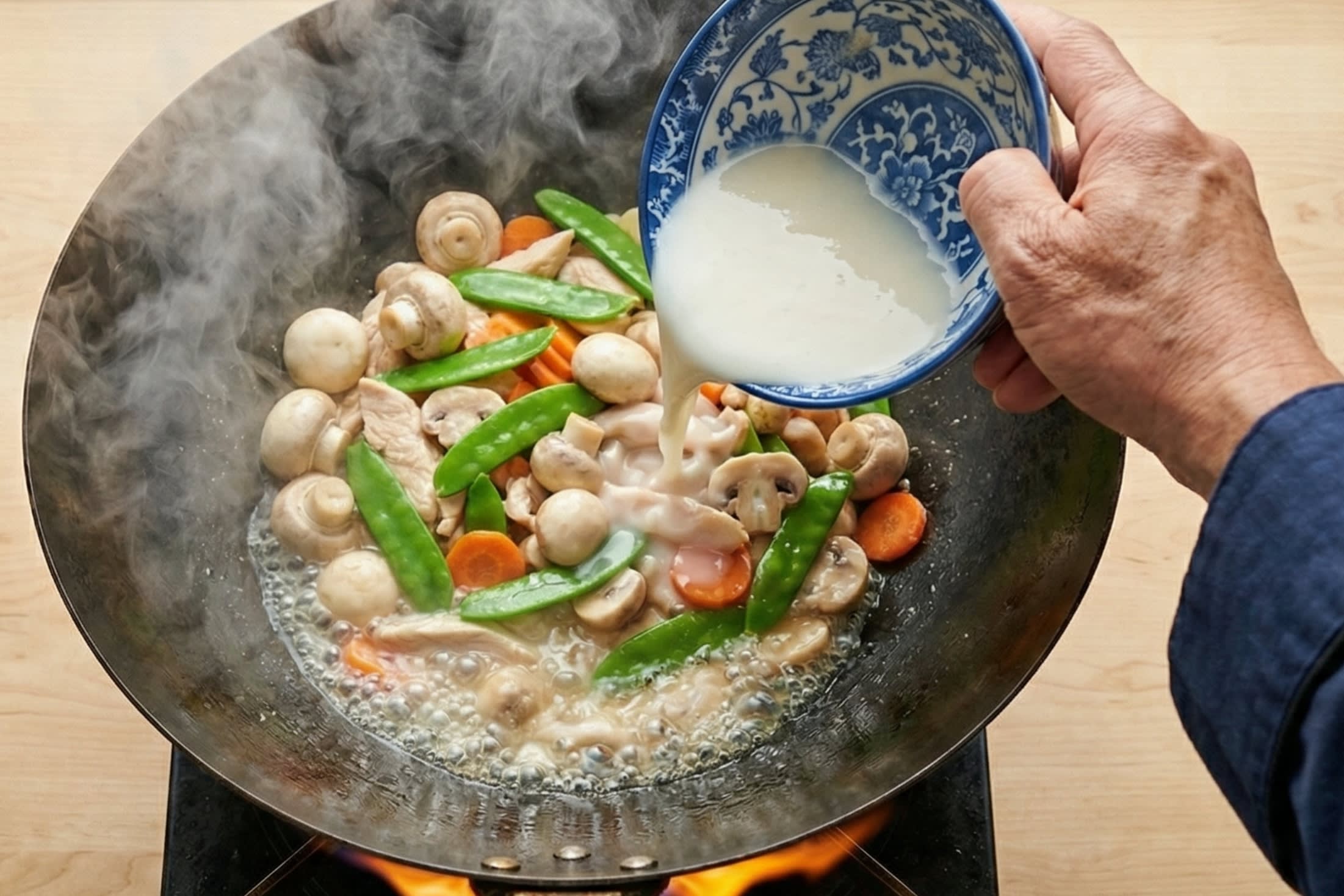 A vibrant action food photography shot looking directly down into a hot, traditional carbon steel wok over a bright flame. A chef is pouring a milky white, highly seasoned chicken broth and cornstarch slurry over a colorful, simmering medley of velvety chicken slices, white mushrooms, bright green snow peas, and carrots. The sauce is visibly bubbling furiously and beginning to thicken into a rich, highly glossy, translucent glaze. Hot steam is rising dramatically.