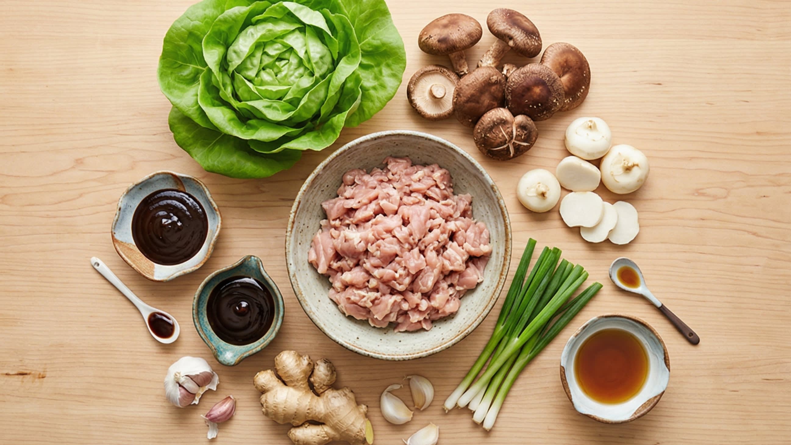 A beautiful flat lay of fresh ingredients on a wooden board: a bowl of hand-minced raw chicken thighs, a whole head of vibrant Butter lettuce, fresh dark shiitake mushrooms, crisp white water chestnuts, garlic cloves, ginger root, scallions, and small ceramic dipping dishes containing thick, dark Hoisin sauce, oyster sauce, and soy sauce.