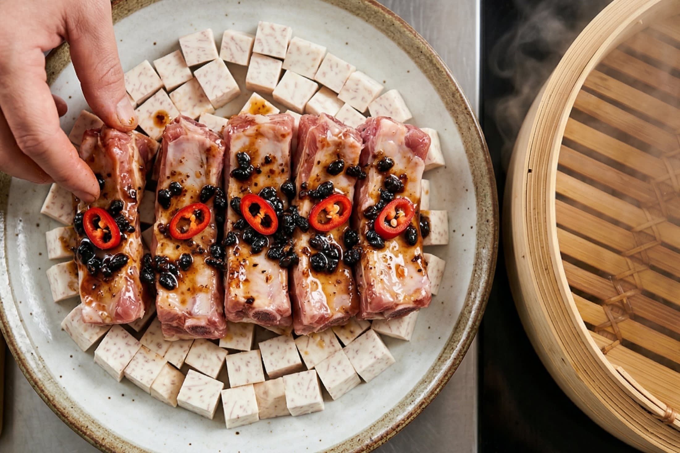 A vibrant food photography shot looking directly down at a rustic ceramic plate. A chef is arranging the sticky, highly glossy, starch-coated spare ribs in a single layer over a bed of pale, cubed taro root. The ribs are speckled with dark black beans, minced garlic, and bright red chili rings. The dish is ready for the steamer.