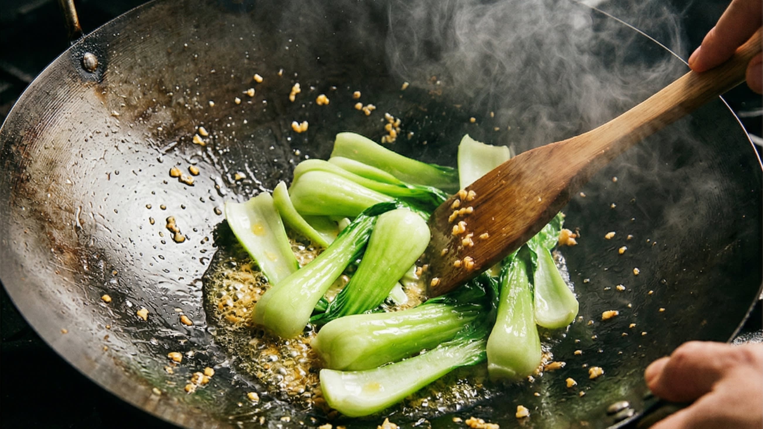 A dynamic close-up action food photography shot looking down into a hot, well-seasoned carbon steel wok. A chef is using a wooden spatula to aggressively stir-fry the thick, pale-green stems of the Baby Bok Choy in sizzling, shimmering garlic oil. The stems are turning a vibrant, bright green. High shutter speed, intense heat, plumes of appetizing steam.