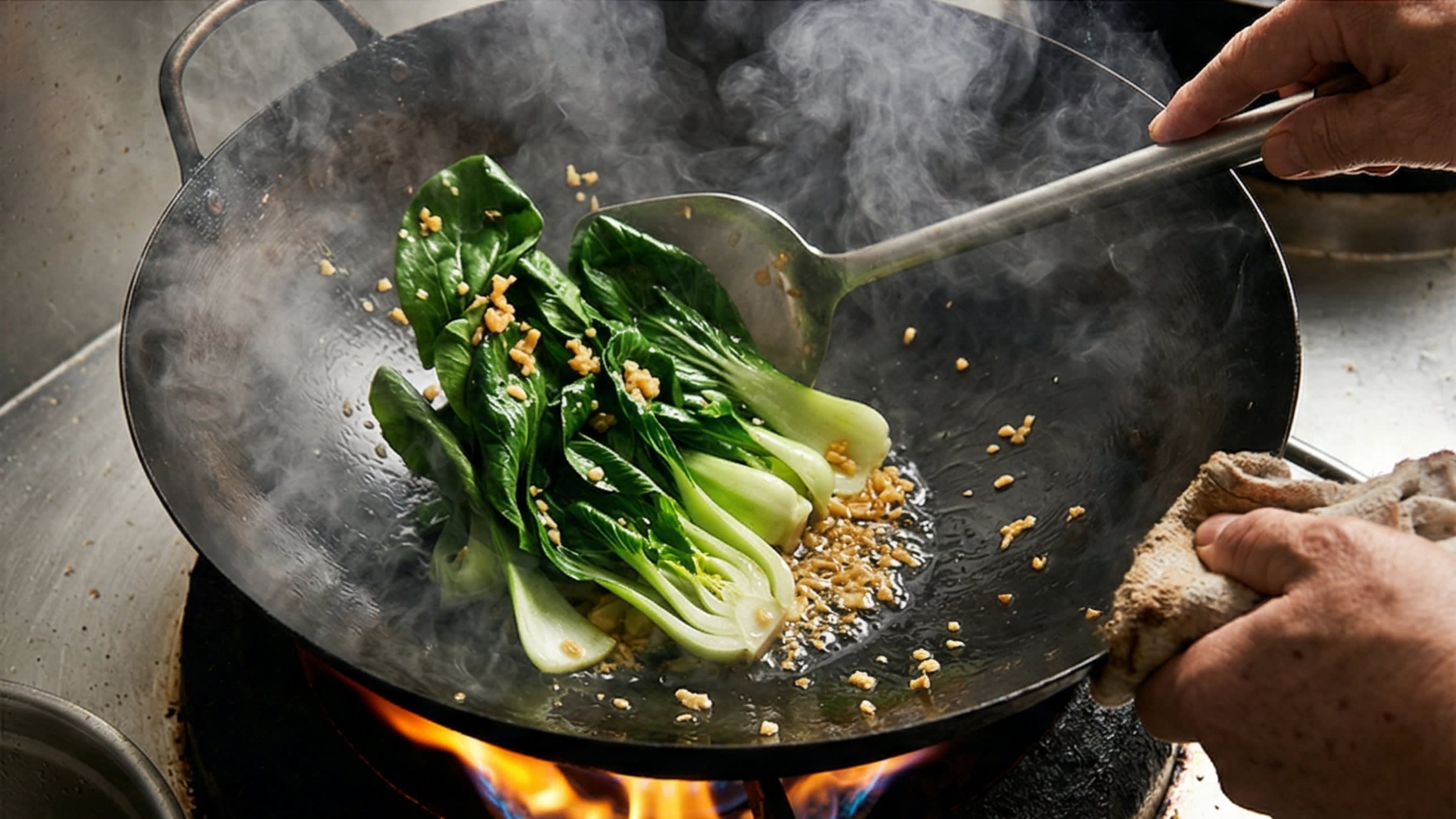 A vibrant action food photography shot looking directly down into a hot, traditional carbon steel wok over a bright roaring flame. A chef is aggressively tossing the wilted, dark green Bok Choy leaves with the crisp pale stems and golden minced garlic. A fine dusting of the seasoning blend is flying through the air. Intense heat, thick plumes of appetizing steam, conveying a sense of explosive wok hei.