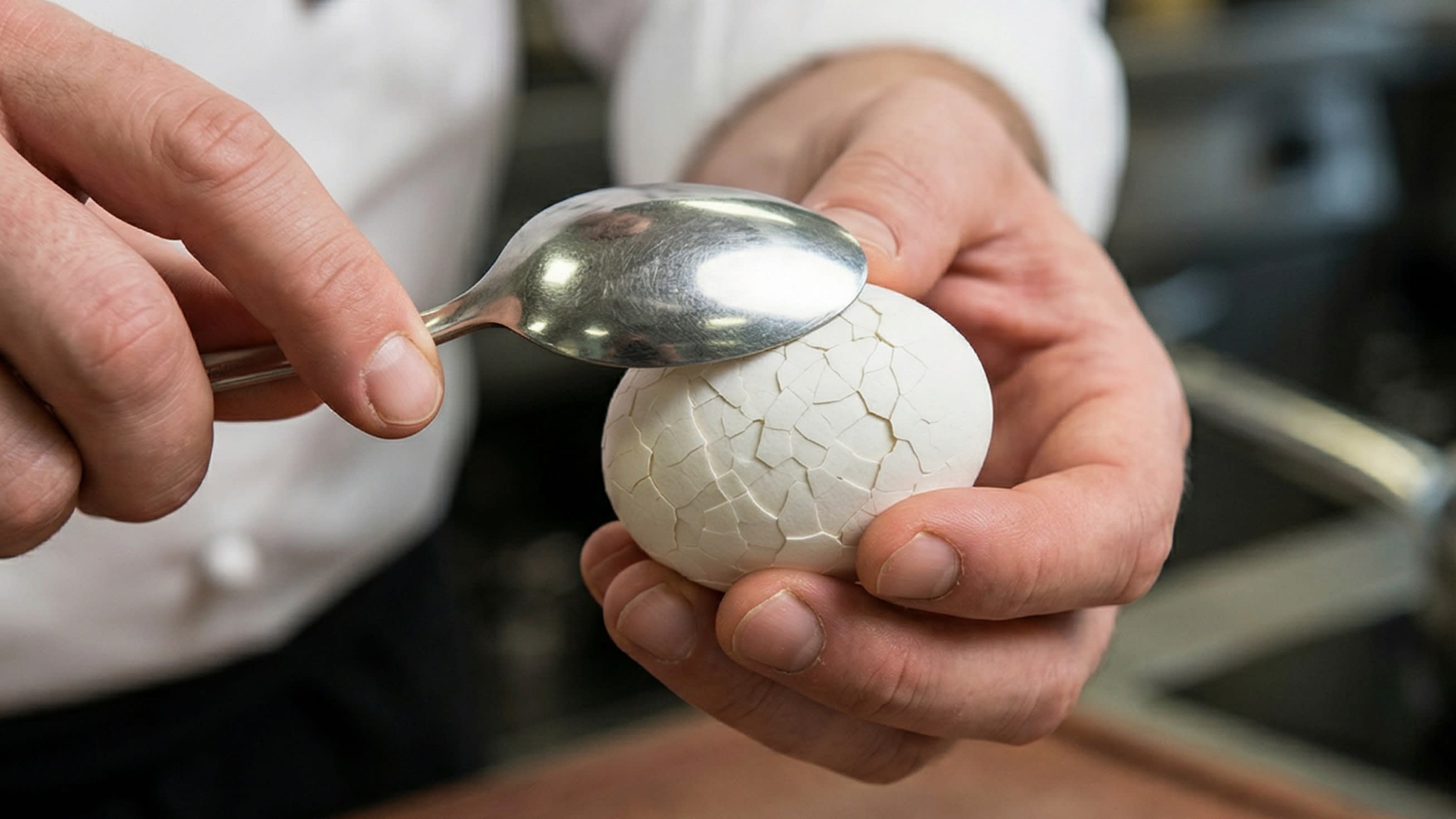 A dynamic close-up action food photography shot focusing on a chef's hands. The chef is holding a boiled egg and gently but firmly tapping the entire surface of the shell with the back of a metal spoon. The shell is visibly fracturing into an intricate, mosaic-like network of tiny cracks without falling off the egg.