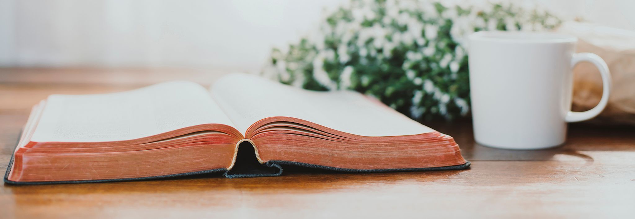 A Bible is sitting on a desk next to a cup of coffee and flowers.