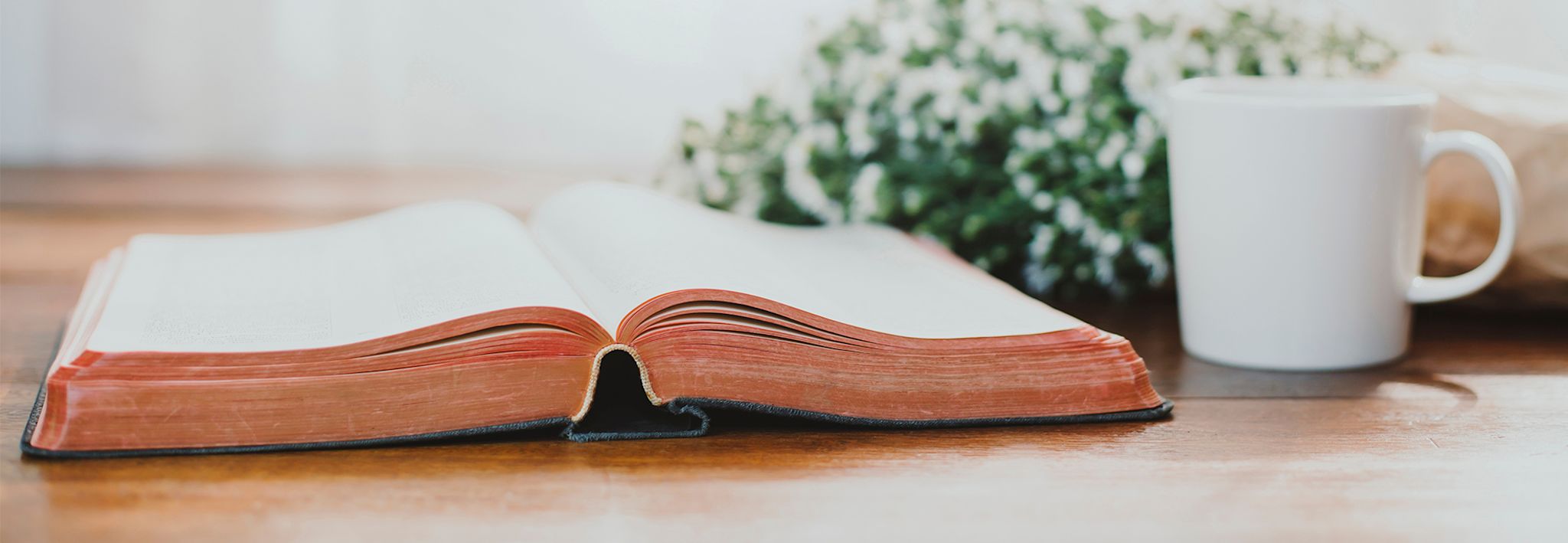 A Bible is sitting on a desk next to a cup of coffee and flowers.