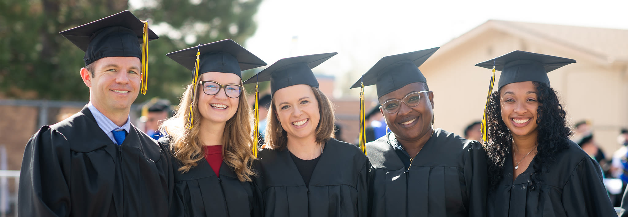Five smiling adult graduate students, dressed in caps and gowns, stand together after completing graduation Five smiling adult graduate students, dressed in caps and gowns, stand together after completing graduation
