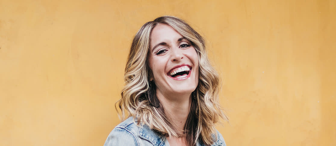 A woman against a yellow background smiling for a photo. (slide 1 of 5)